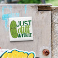 A sticker with a cartoon pickle and the text "Just dill with it" is attached to a concrete surface.