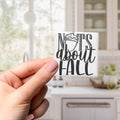 A hand holding a small white sticker with the text "NOTS ABOUT FALL" printed on it.