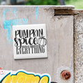 a white square tile with the text "Pumpkin spice everything" written on it, which is attached to a concrete wall.