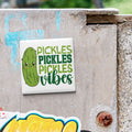 A sticker with a cartoon pickle and the text "Pickles, Pickles, Vibes" is attached to a concrete surface.