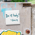 a white square magnet with the text "Do it lady! - Chit" written on it, attached to a weathered concrete surface.