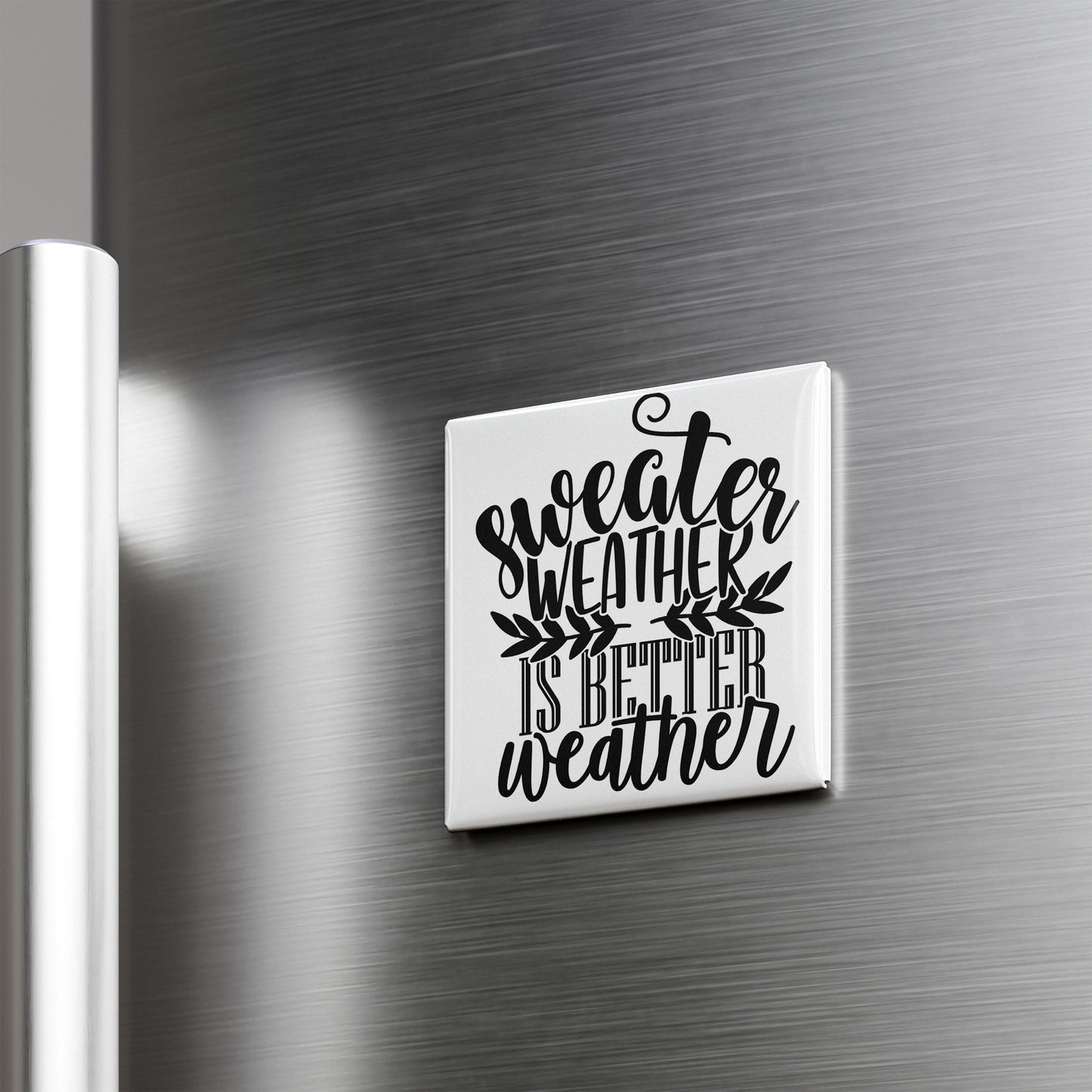 A white square magnet with the text " sweater weather is better weather" printed on it, mounted on a metallic surface.