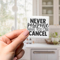 A hand holding a white sticker with the text "NEVER postone what you can cancel" printed on it.