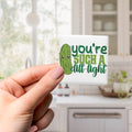 A person's hand holding a square magnet with a cartoon image of a pickle and the text "you're such a dill-tight" printed on it.