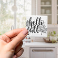 A hand holding a small square tile with the text "Hello fall" written on it.