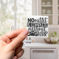 A person's hand holding a sticker with the text "NO OUTFIT IS COMPLETE without GHAIR" against a blurred background of a kitchen.