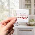 A hand holding a small square magnet with the text "kindness is magic" written on it.