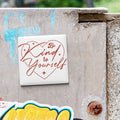 A white tile with the text "Be Kind to Yourself" in red cursive lettering is attached to a concrete wall.
