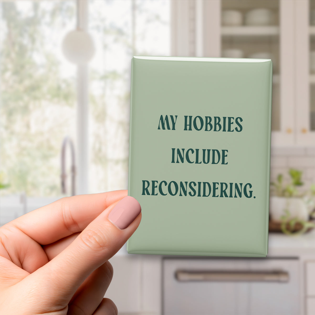 Hand holding the 2.5 × 3.5-inch “My hobbies include reconsidering.” magnet in a kitchen