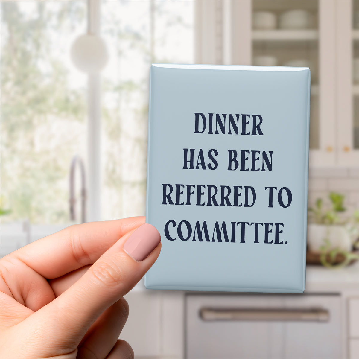 Hand holding the 2.5 × 3.5-inch “Dinner has been referred to committee.” magnet in a kitchen