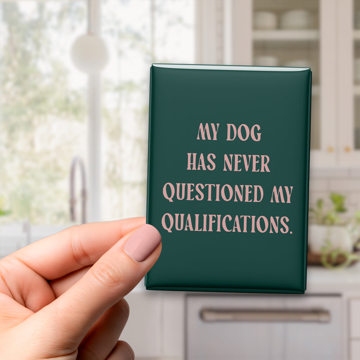 Hand holding the 2.5 × 3.5-inch “My dog has never questioned my qualifications.” magnet in a kitchen