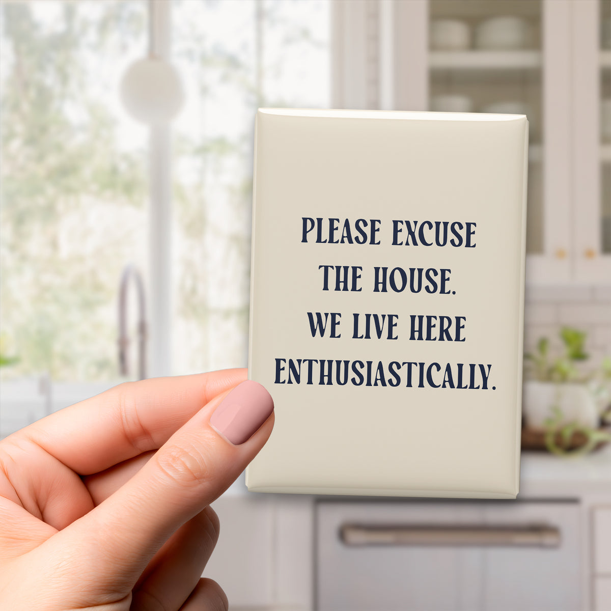 Hand holding the 2.5 × 3.5-inch “Please excuse the house. We live here enthusiastically.” magnet in a kitchen