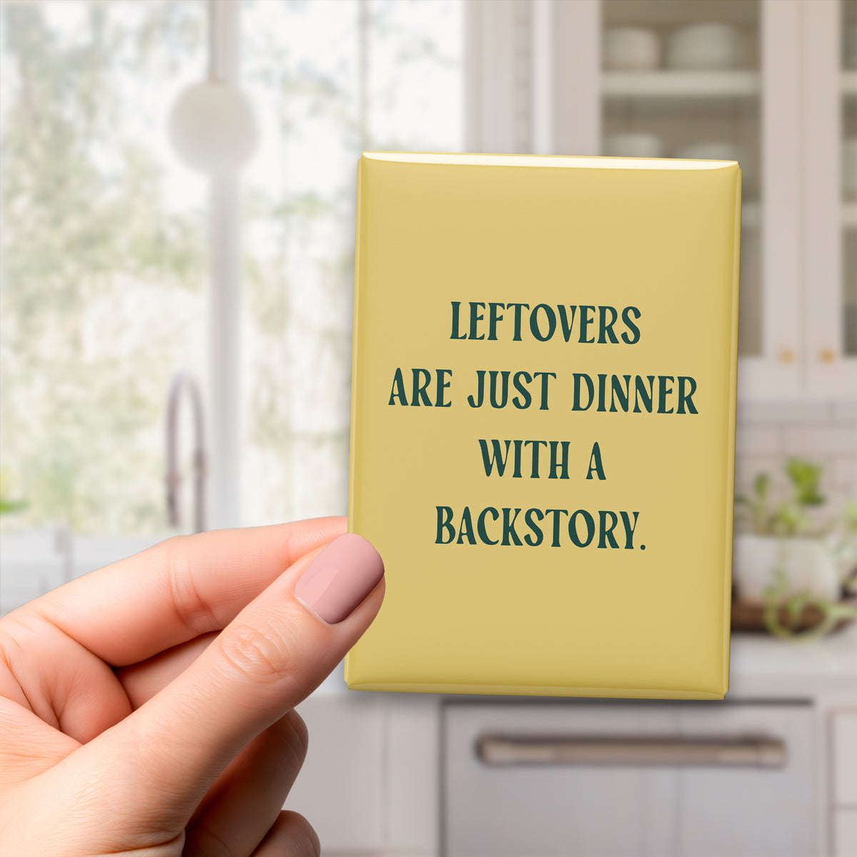 Hand holding the 2.5 × 3.5-inch “Leftovers are just dinner with a backstory.” magnet in a kitchen
