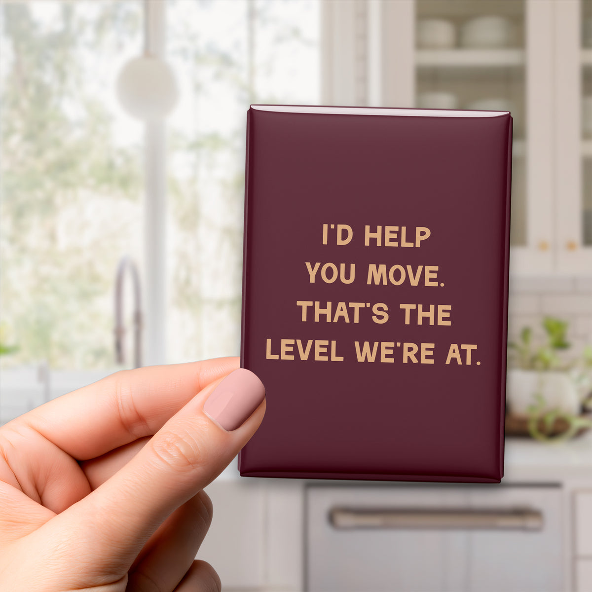 Hand holding the 2.5 × 3.5-inch “I'd help you move. That's the level we're at.” magnet in a kitchen