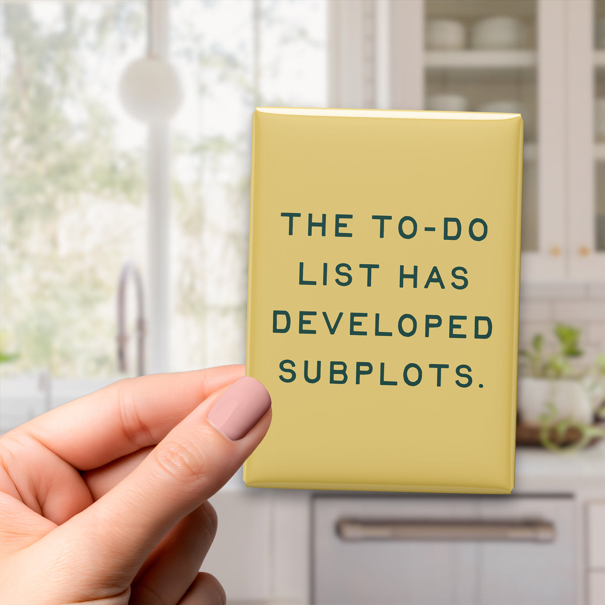 Hand holding the 2.5 × 3.5-inch “The to-do list has developed subplots.” magnet in a kitchen