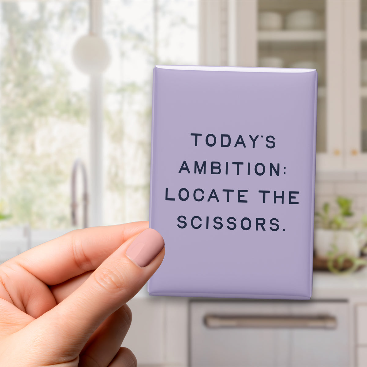 Hand holding the 2.5 × 3.5-inch “Today's ambition: locate the scissors.” magnet in a kitchen