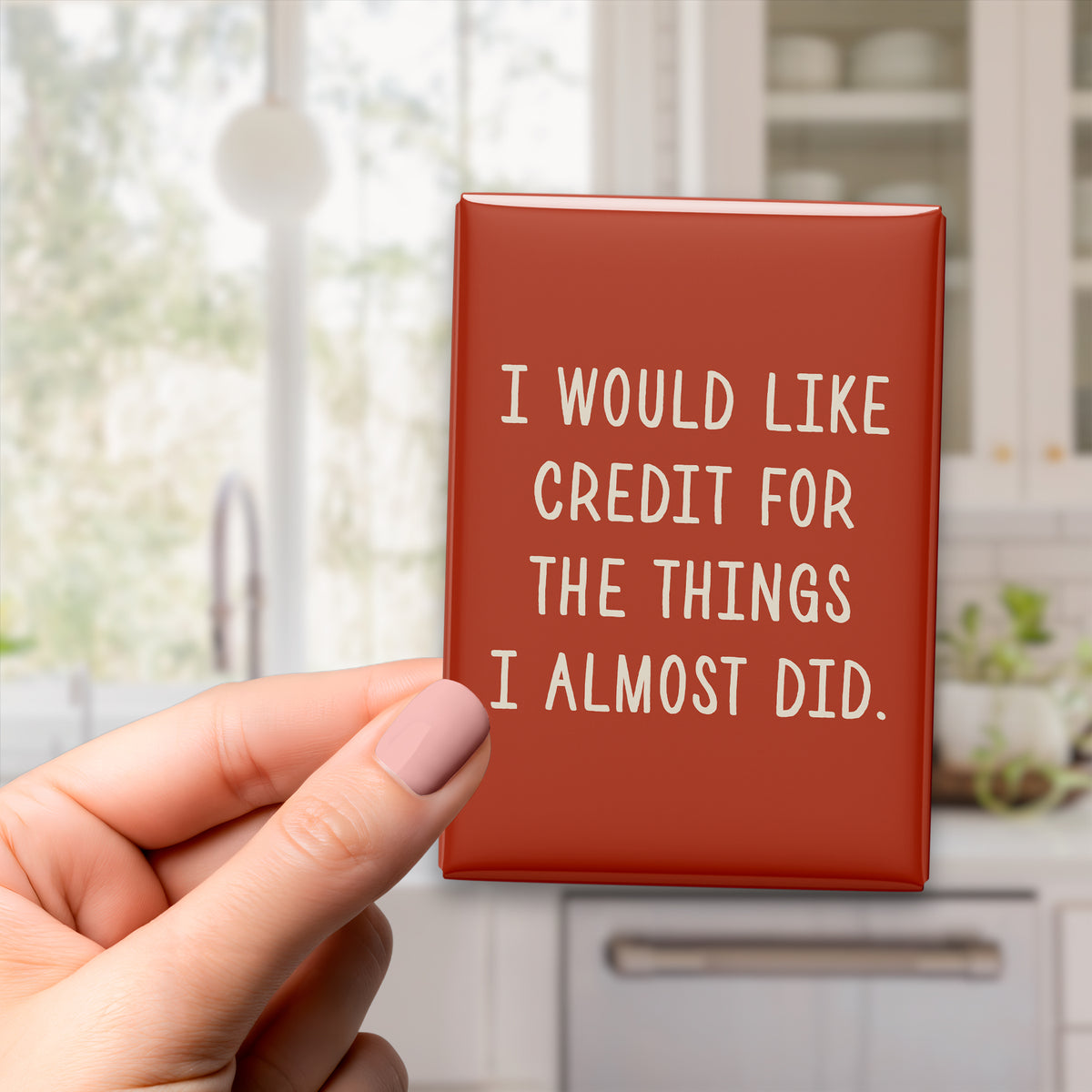 Hand holding the 2.5 × 3.5-inch “I would like credit for the things I almost did.” magnet in a kitchen