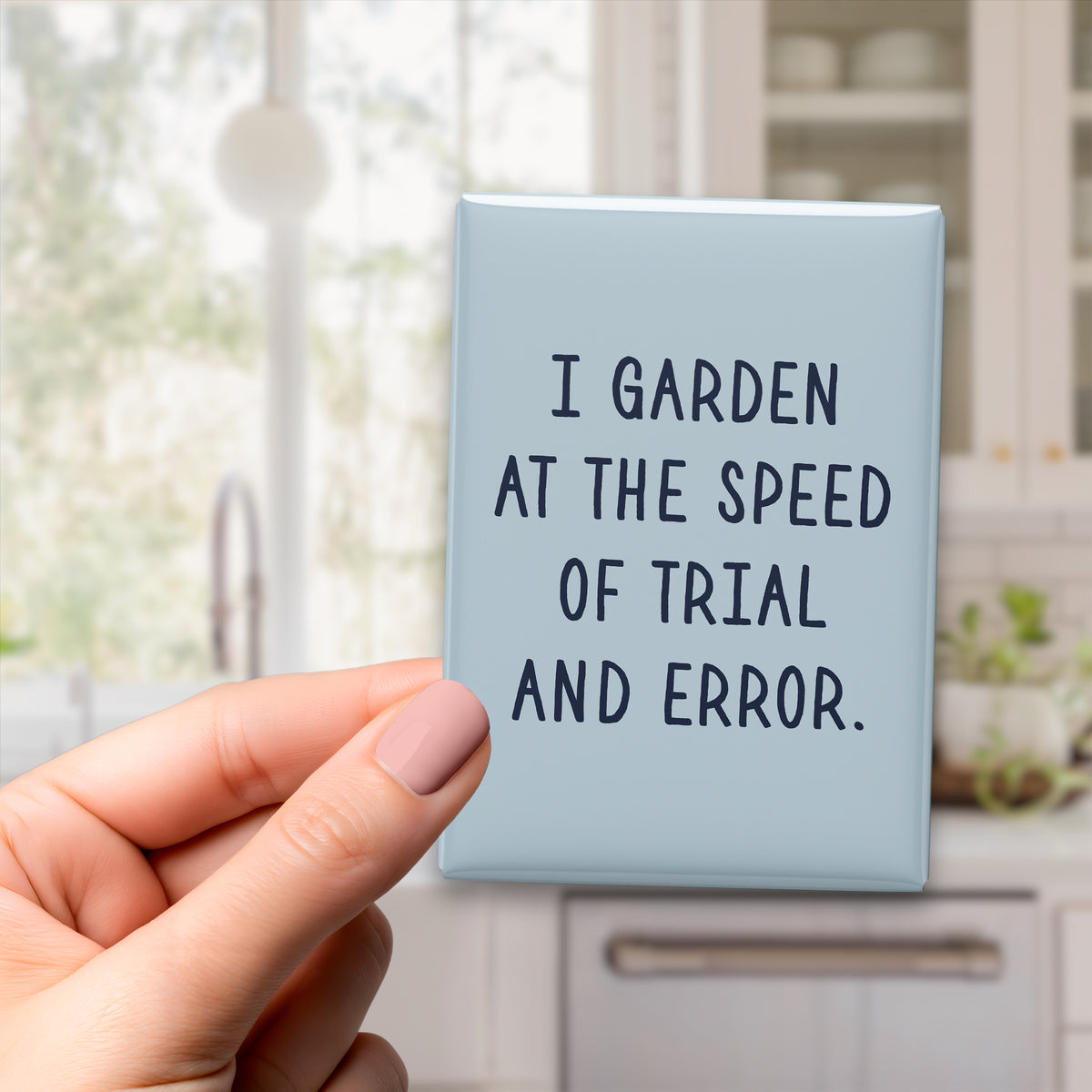 Hand holding the 2.5 × 3.5-inch “I garden at the speed of trial and error.” magnet in a kitchen