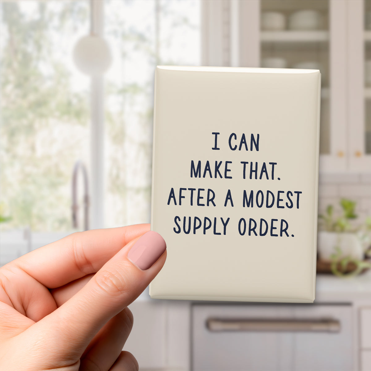 Hand holding the 2.5 × 3.5-inch “I can make that. After a modest supply order.” magnet in a kitchen