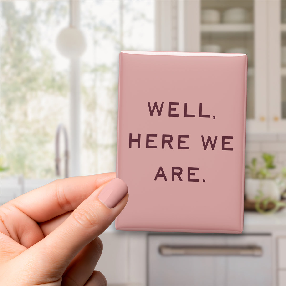 Hand holding the 2.5 × 3.5-inch “Well, here we are.” magnet in a kitchen