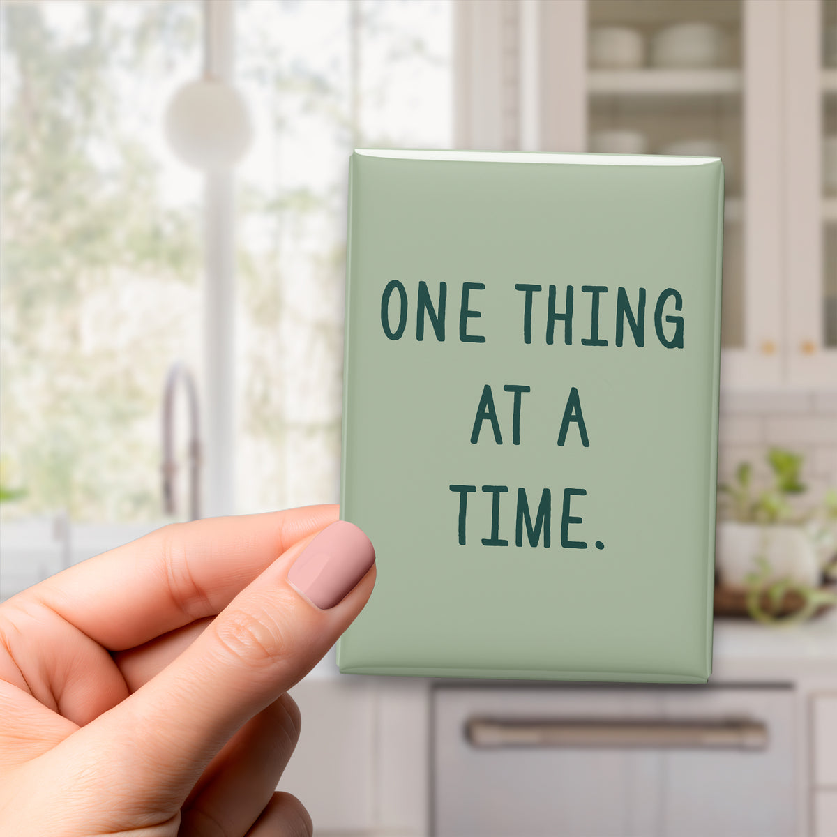 Hand holding the 2.5 × 3.5-inch “One thing at a time.” magnet in a kitchen