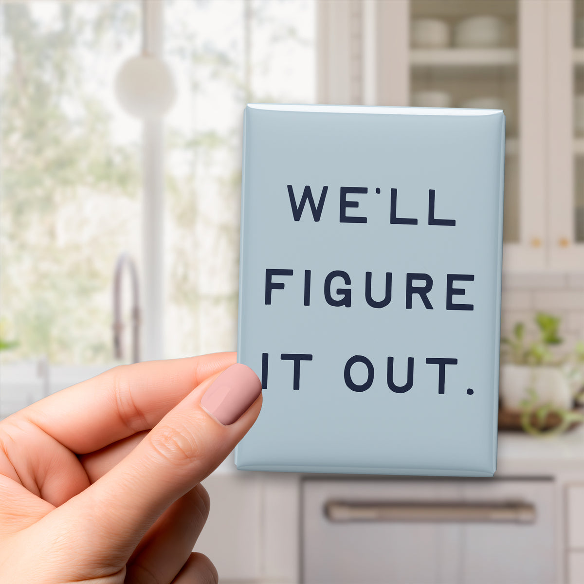 Hand holding the 2.5 × 3.5-inch “We'll figure it out.” magnet in a kitchen