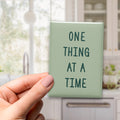 One Thing at a Time magnet in Spruce lettering on a Sage background, held in a hand in a kitchen