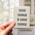 Good Enough Is Good Enough magnet in Midnight lettering on a Parchment background, held in a hand in a kitchen