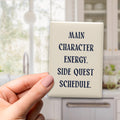 Main Character Energy. Side Quest Schedule magnet in Midnight lettering on a Parchment background, held in a hand in a kitchen