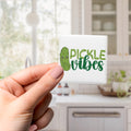A hand holding a small square magnet with the text "Pickle Vibes" on it.