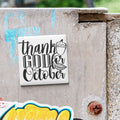 a white square magnet with black text that says "Thank GOD October" attached to a concrete wall.