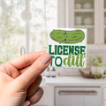 A hand holding a sticker with a cartoon pickle and the text "License to dill" printed on it.