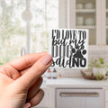 A hand holding a sticker with the text "I'd love to buy my dog said no" against a blurred background of a kitchen.
