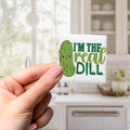 A person's hand holding a small square magnet with a cartoon image of a pickle and the text "I'm the real dill" printed on it.