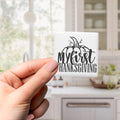 A hand holding a small white square with a black text design that says "My first Thanksgiving".