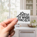 A hand holding a small square magnet with the text "home is where the cat is" written on it.