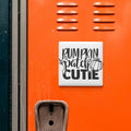 a close-up of an orange surface with a white sign that reads "Pumpkin Patch Cutie" in black text.