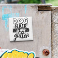 A white tile with the text "DOG HAIR IS MY GLITTER" is attached to a concrete wall.