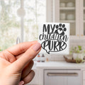 A hand holding a small square magnet with the text "My children purr" on it.