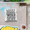 A white tile with black text that says "GIVE thanks with a grateful heart" is attached to a concrete wall.