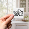 A hand holding a square magnet with the text "sweater weather" written on it.