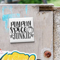 a white square tile with the text "Pumpkin spice junkie" written on it, which is attached to a concrete wall.