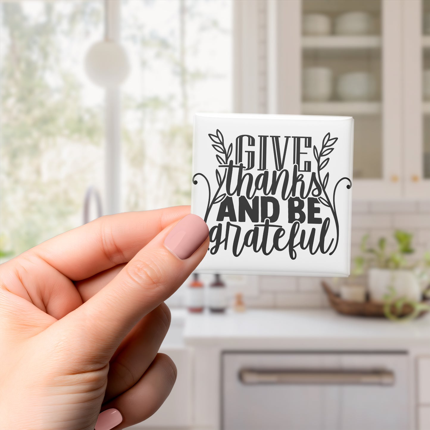 A hand holding a small square magnet with the text "GIVE thanks AND BE grateful" written on it.