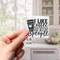A hand holding a sticker with the text "I LIKE coffee AND maybe 3 people" against a blurred background of a kitchen.