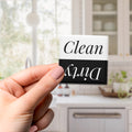 A hand holding a black and white sign with the text "Clean" and "Dirty" on it, with a blurred background of a kitchen counter and cabinets.