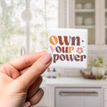 A hand holding a square magnet with the text "own your power" written on it.