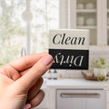 A hand holding a black and white sign with the text "Clean" and "Dirty" on it, with a blurred background of a kitchen counter and cabinets.