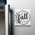 A refrigerator door with a decorative magnet that says "fall" surrounded by a leaf and a sunburst design.