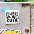 a white square tile with the text "Rumpkin Patch Cutie" written on it, which is attached to a concrete wall.