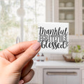 A hand holding a small square magnet with the text "Thankful Gratified Blessed" written on it.